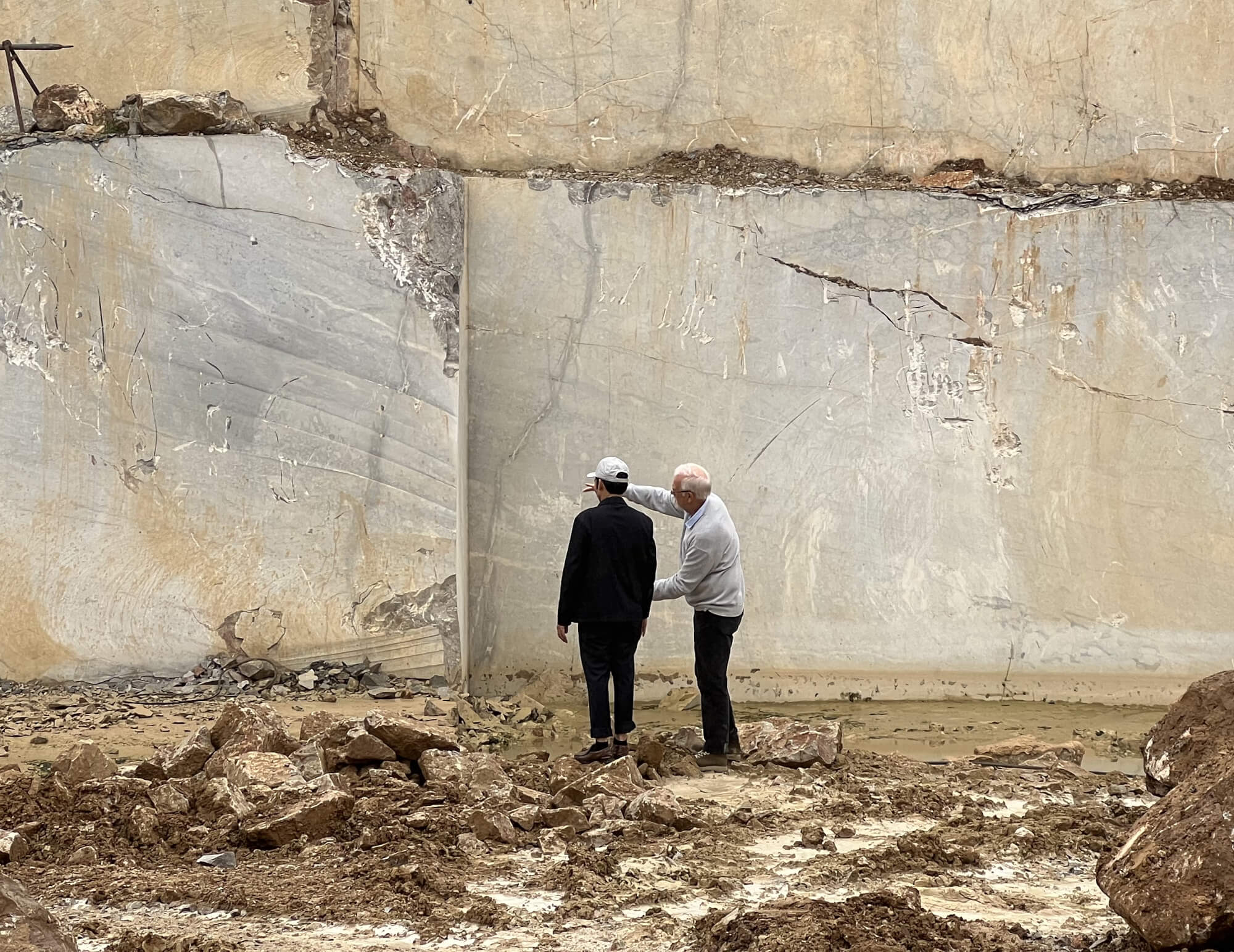 Two men standing in the middle of a quarry, facing a large rock wall, with one man gesturing to the other.